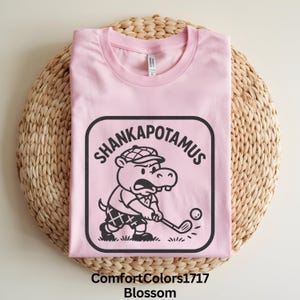 Puede incluir: Camiseta rosa claro con un gráfico de un hipopótamo jugando al golf, con la etiqueta "Shankapotamus". El hipopótamo lleva gorra de golf y pantalones cortos a cuadros. La camiseta está etiquetada como "ComfortColors1717 Blossom".