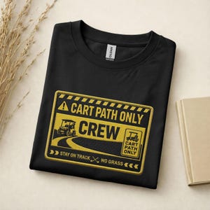 Könnte beinhalten: Schwarzes T-Shirt mit einem gelb-schwarzen Aufnäher mit der Aufschrift "A CART PATH ONLY CREW". Der Aufnäher zeigt eine Golfwagen-Grafik und den Text "STAY ON TRACK, NO GRASS". Das Shirt hat einen Rundhalsausschnitt und ein Gildan-Etikett.