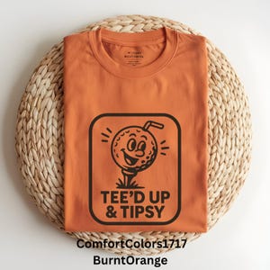 Puede incluir: Camiseta naranja quemado con un gráfico de una bola de golf sonriente en un tee, con el texto "TEE'D UP & TIPSY". La camiseta está sobre una estera circular tejida. La camiseta es un ComfortColors1717.