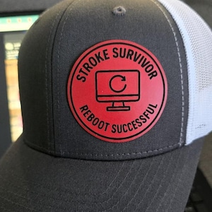 May include: A gray trucker hat with a white mesh back and a red circular patch. The patch features the words "STROKE SURVIVOR" and "REBOOT SUCCESSFUL" around a computer monitor icon with a circular arrow.