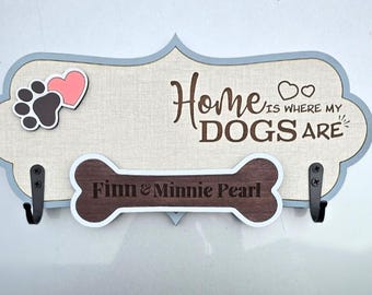 Könnte beinhalten: Ein Holzschild mit einem knochenförmigen Haken und dem Text "Home is where my DOGS ARE" mit den Namen "Finn & Minnie Pearl" auf dem Knochen. Das Schild ist blau lackiert und hat einen hellbraunen Stoffhintergrund.