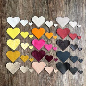 Puede incluir: Una colección de recortes en forma de corazón en varios tamaños y colores, incluyendo blanco, amarillo, naranja, rosa, rojo, negro y beige. Los corazones están dispuestos sobre una superficie de madera, creando una exhibición visualmente atractiva.