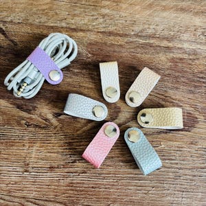 Op de afbeelding: Een verzameling van zeven kleine kabelorganizers gemaakt van getextureerd materiaal in diverse pastelkleuren, waaronder lavendel, wit, beige, lichtgrijs, geel, roze en blauw, elk voorzien van een ronde zilveren metalen drukknoopsluiting.