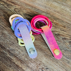 May include: Two hair tie organizers featuring a snap closure. One organizer has a purple and blue gradient glitter finish with a silver snap, holding pastel hair ties. The other has a pink and orange iridescent finish with a gold snap, holding neon hair ties.