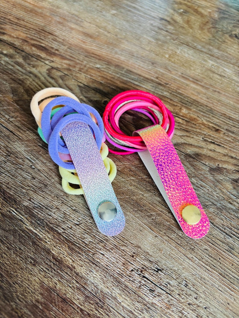 May include: Two hair tie organizers featuring a snap closure. One organizer has a purple and blue gradient glitter finish with a silver snap, holding pastel hair ties. The other has a pink and orange iridescent finish with a gold snap, holding neon hair ties.