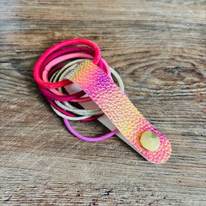 May include: A collection of elastic hair ties in shades of hot pink, light pink, silver, and purple, held together by a textured, iridescent pink and gold faux leather strap secured with a single round gold-toned metal snap button fastener.