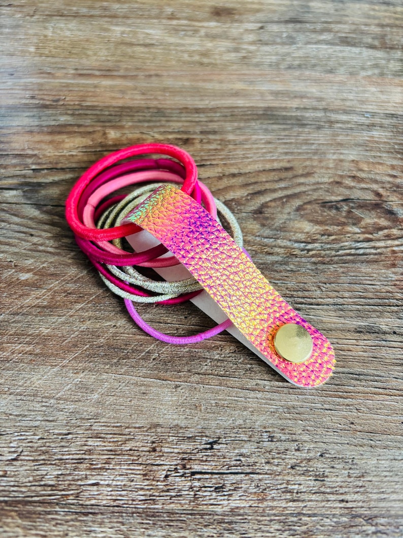 May include: A collection of elastic hair ties in shades of hot pink, light pink, silver, and purple, held together by a textured, iridescent pink and gold faux leather strap secured with a single round gold-toned metal snap button fastener.