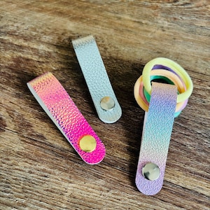 May include: Three rectangular folded straps with metal snap buttons. One strap is silver, one is pink and orange iridescent, and one is a blue and purple gradient holding a cluster of pastel-colored elastic hair ties in yellow, peach, and green.