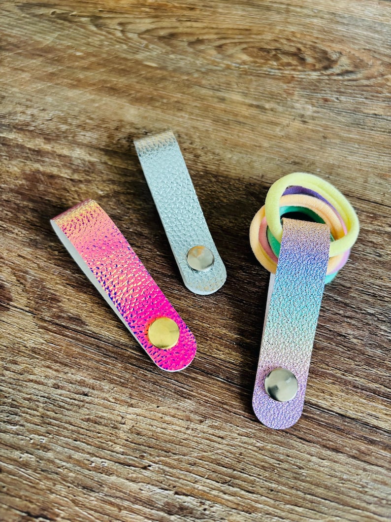 May include: Three rectangular folded straps with metal snap buttons. One strap is silver, one is pink and orange iridescent, and one is a blue and purple gradient holding a cluster of pastel-colored elastic hair ties in yellow, peach, and green.