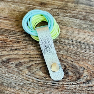 May include: A silver textured leather strap with a metallic snap button closure holding a stack of light blue and neon yellow elastic hair ties. The strap is folded over the circular hair ties to keep them organized in a single bundle.