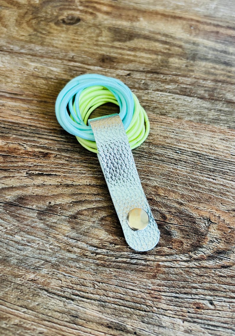 May include: A silver textured leather strap with a metallic snap button closure holding a stack of light blue and neon yellow elastic hair ties. The strap is folded over the circular hair ties to keep them organized in a single bundle.