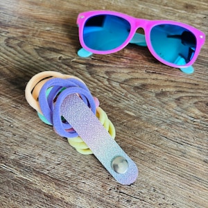 May include: Bright pink sunglasses with reflective blue lenses positioned above a collection of pastel-colored elastic hair ties held together by a glittery, ombre-toned fabric strap featuring a single silver-colored metal snap button closure.