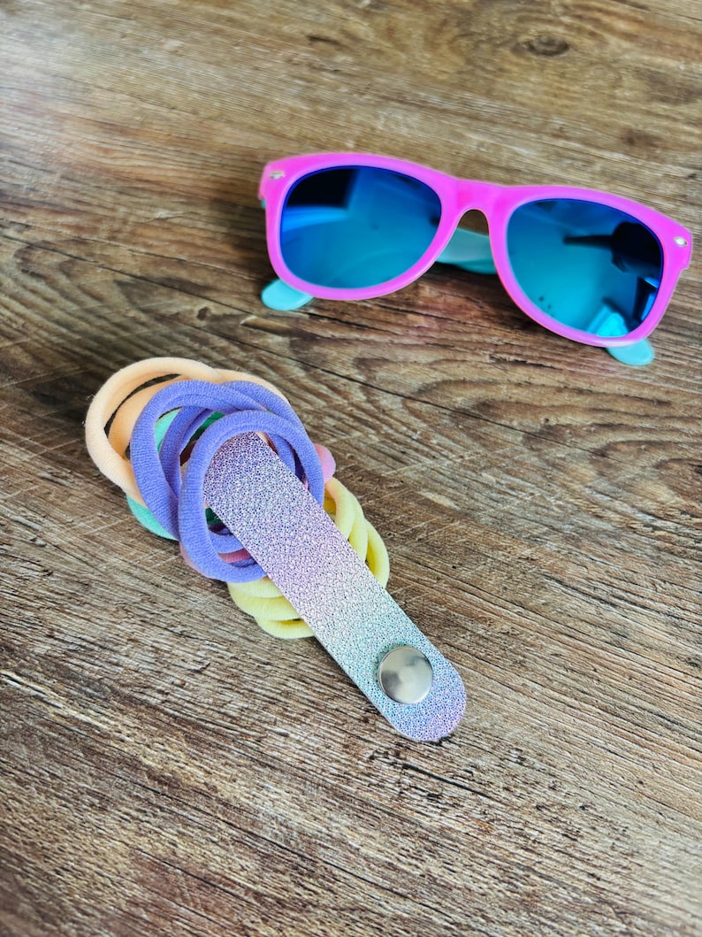 May include: Bright pink sunglasses with reflective blue lenses positioned above a collection of pastel-colored elastic hair ties held together by a glittery, ombre-toned fabric strap featuring a single silver-colored metal snap button closure.