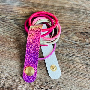 May include: A hair tie holder featuring a strap with a vibrant, iridescent purple and pink textured finish. The strap is secured with a gold-toned metal snap button and holds a collection of elastic hair bands in shades of pink, magenta, and gold.