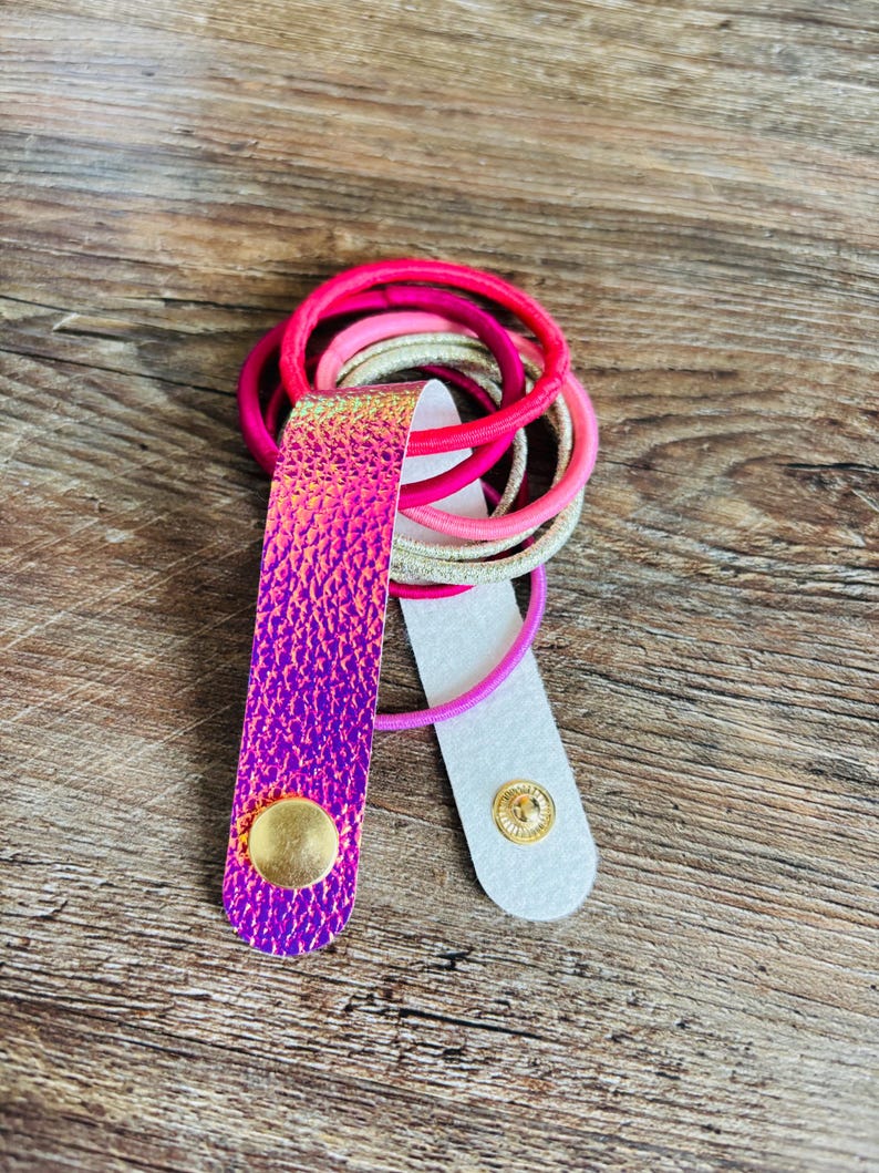 May include: A hair tie holder featuring a strap with a vibrant, iridescent purple and pink textured finish. The strap is secured with a gold-toned metal snap button and holds a collection of elastic hair bands in shades of pink, magenta, and gold.