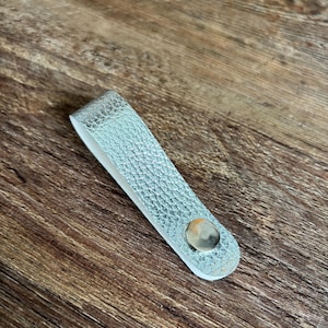 May include: A silver-colored, textured loop handle with a metallic finish, featuring a single polished silver-tone metal rivet fastener at the base. The object is folded into a U-shape, displaying a smooth white interior lining along the inner edge.