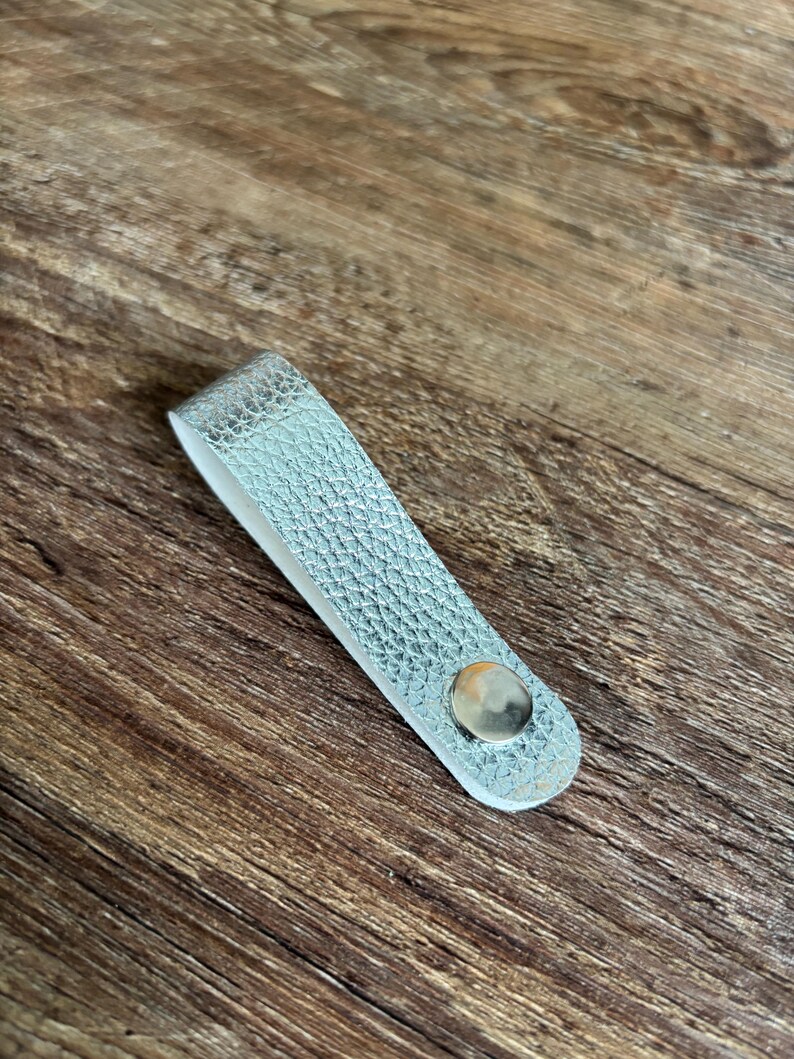 May include: A silver-colored, textured loop handle with a metallic finish, featuring a single polished silver-tone metal rivet fastener at the base. The object is folded into a U-shape, displaying a smooth white interior lining along the inner edge.