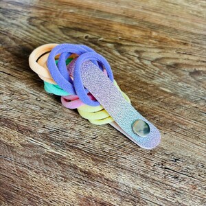 May include: A collection of elastic hair ties in pastel shades of peach, mint green, pink, and yellow, held together by a single, elongated, glittery purple-to-blue gradient strap secured with a small, circular, silver-toned metal snap button.
