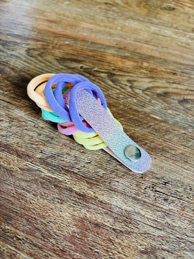 May include: A collection of elastic hair ties in pastel shades of peach, mint green, pink, and yellow, held together by a single, elongated, glittery purple-to-blue gradient strap secured with a small, circular, silver-toned metal snap button.