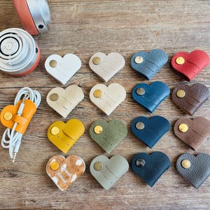 Könnte beinhalten: Eine Sammlung von herzförmigen Kabelorganisatoren aus Leder in verschiedenen Farben wie Weiß, Beige, Rot, Blau, Gelb, Grün, Braun und Grau, jeweils mit einem metallischen Druckknopfverschluss, neben einem Paar Over-Ear-Kopfhörern.