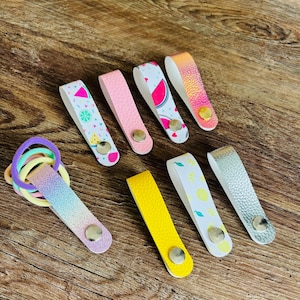 May include: A collection of seven faux leather hair tie holders with metal snap buttons. The holders feature various designs including watermelon prints, solid pink, solid yellow, iridescent, silver, and lemon patterns, holding colorful hair ties.
