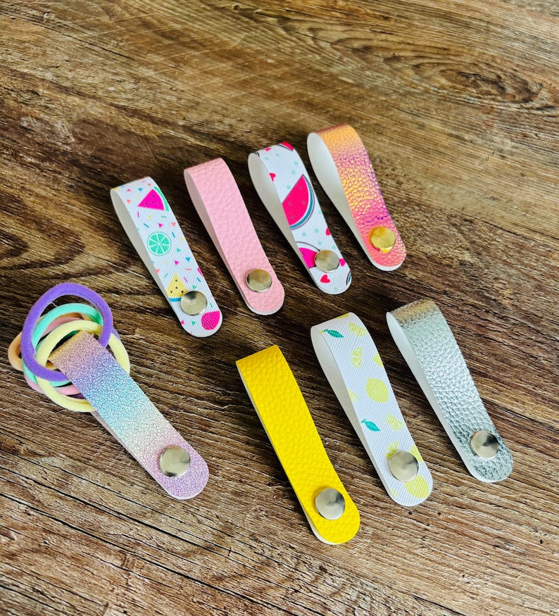 May include: A collection of seven faux leather hair tie holders with metal snap buttons. The holders feature various designs including watermelon prints, solid pink, solid yellow, iridescent, silver, and lemon patterns, holding colorful hair ties.