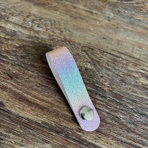 May include: A folded strap with a textured, iridescent finish that shifts between shades of pink, blue, and purple. The strap is secured at the bottom with a circular, polished silver-toned metal snap button, creating a loop shape.
