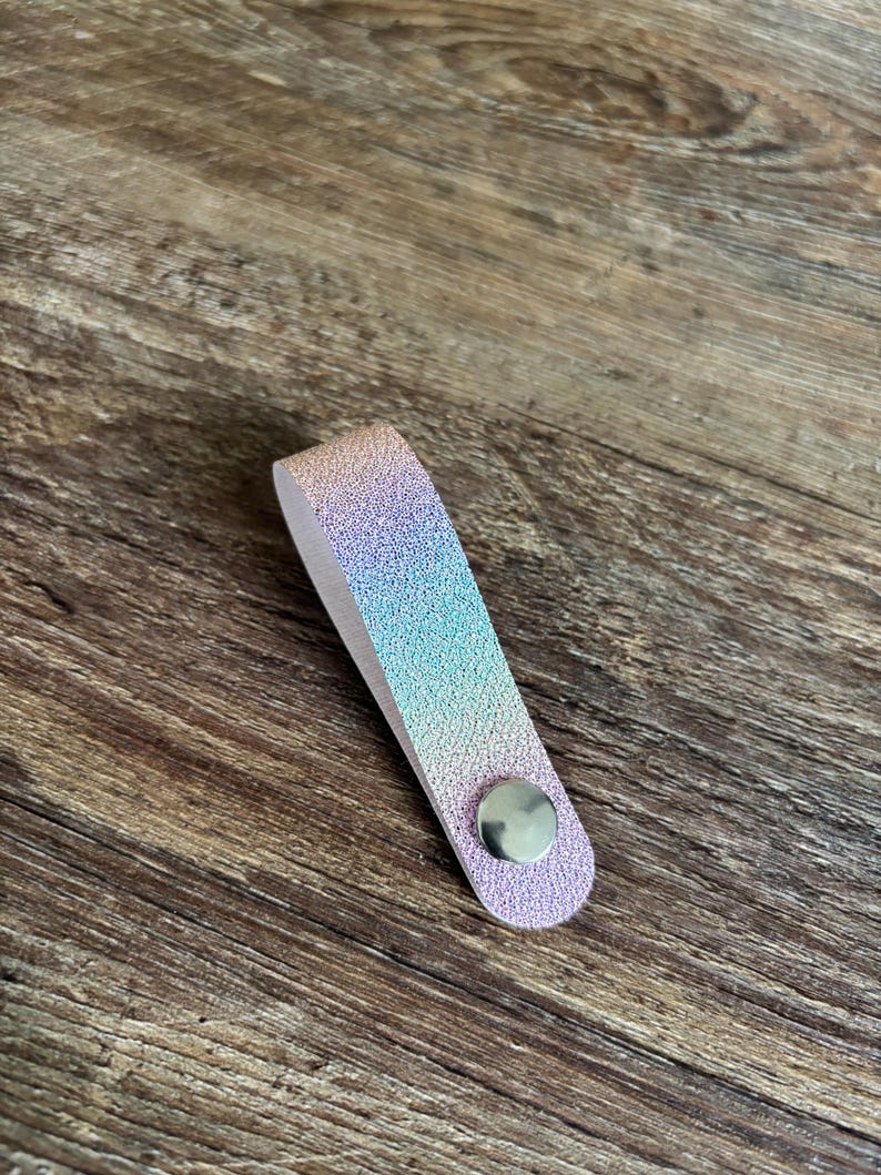 May include: A folded strap with a textured, iridescent finish that shifts between shades of pink, blue, and purple. The strap is secured at the bottom with a circular, polished silver-toned metal snap button, creating a loop shape.