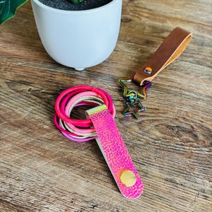 May include: A set of hair ties in shades of pink and gold held by a bright pink holographic strap with a gold snap button, alongside a brown leather keychain featuring a rainbow-colored metal star-shaped clasp and a smaller star charm.