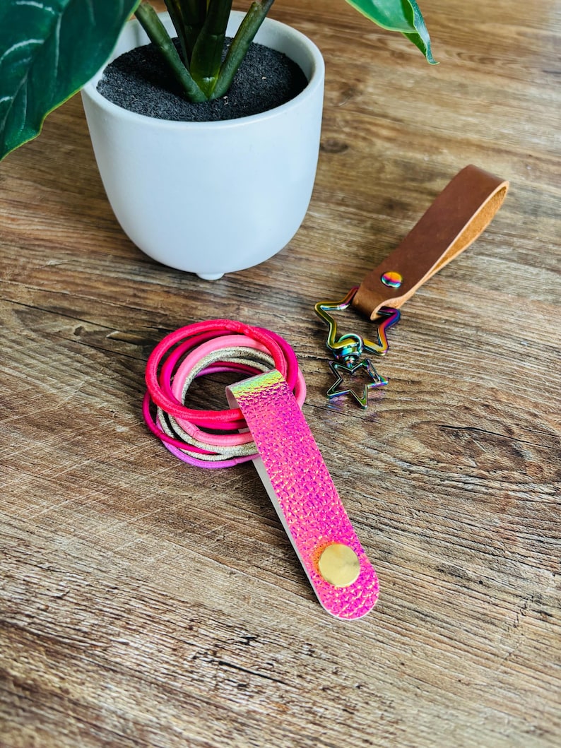 May include: A set of hair ties in shades of pink and gold held by a bright pink holographic strap with a gold snap button, alongside a brown leather keychain featuring a rainbow-colored metal star-shaped clasp and a smaller star charm.