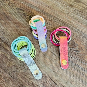 May include: Three hair tie organizers featuring metallic and iridescent faux leather straps with snap buttons. One strap is silver, one is a purple-to-blue gradient, and one is a pink-to-orange iridescent finish, each holding multiple elastic hair ties.