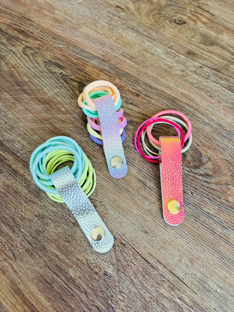 May include: Three hair tie organizers featuring metallic and iridescent faux leather straps with snap buttons. One strap is silver, one is a purple-to-blue gradient, and one is a pink-to-orange iridescent finish, each holding multiple elastic hair ties.