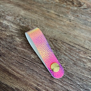 May include: A folded loop strap featuring a vibrant iridescent pink and gold textured surface. The strap is secured at the bottom with a circular, smooth, metallic gold-toned fastener, creating a clean and functional design element.