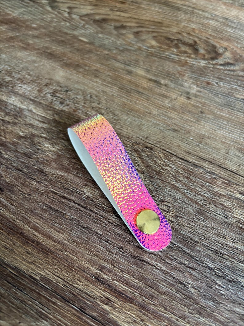 May include: A folded loop strap featuring a vibrant iridescent pink and gold textured surface. The strap is secured at the bottom with a circular, smooth, metallic gold-toned fastener, creating a clean and functional design element.