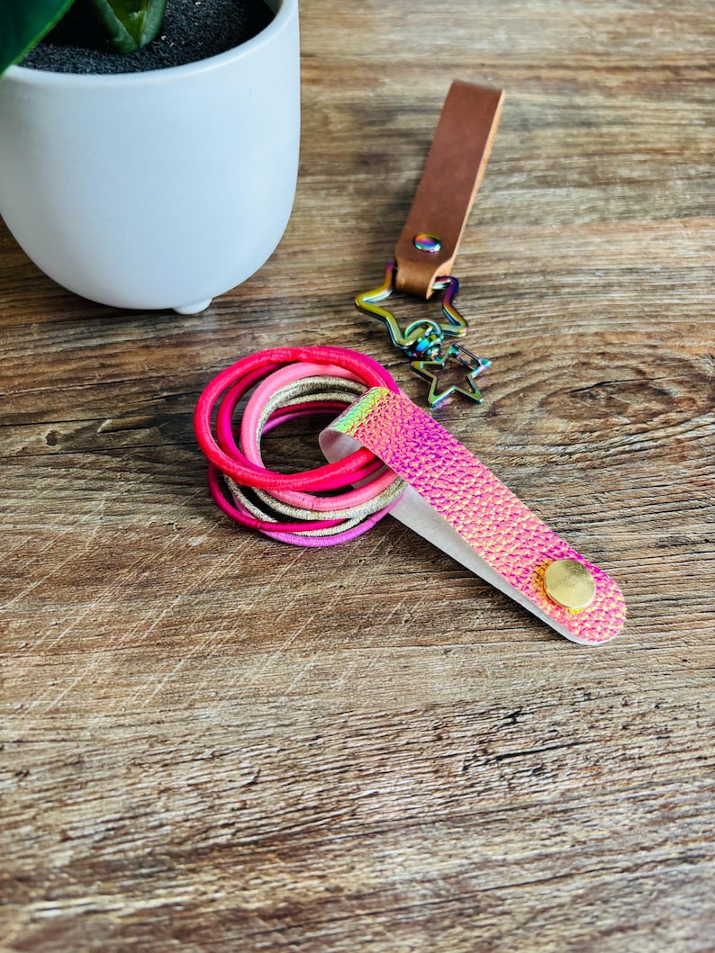 May include: A set of elastic hair ties in shades of pink and gold, held together by a textured, iridescent pink strap with a gold-tone snap button. A brown leather strap with a rainbow-colored metal star-shaped clip is attached to the hair ties.