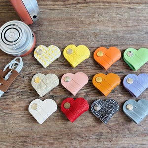 Puede incluir: Una colección de doce organizadores de cables en forma de corazón de varios colores y texturas, incluyendo amarillo, naranja, verde, rosa, azul, rojo, blanco y negro, cada uno con un cierre de botón metálico, junto a unos auriculares.