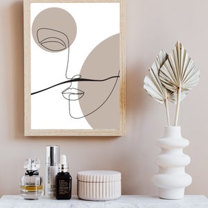 May include: A framed art print with a minimalist line drawing of a face in black against a white and beige backdrop. The artwork is in a light wood frame. A vase with dried leaves and other decorative items are on a shelf below the artwork.