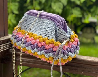 May include: A colorful, crocheted clutch with a silver chain strap. The bag features a mix of colors, including blue, purple, pink, and yellow. The bag is resting on a wooden railing, with a green background.