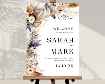 May include: A wedding welcome sign on a wooden easel. The sign features floral watercolor artwork in neutral tones and the names "SARAH" and "MARK". The text reads "WELCOME TO THE WEDDING OF" and the date "16.10.2X".