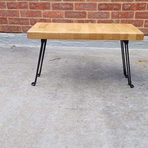 May include: A rectangular wooden coffee table with a light brown surface. The table has four black metal hairpin legs. The background features a red brick wall and a concrete surface.