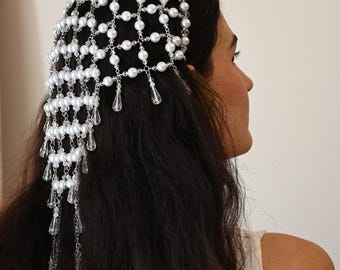 May include: A headpiece featuring a delicate arrangement of white faux pearls and clear crystal teardrop pendants. The headpiece is designed with a net-like structure, with the pearls connected by silver-toned chains. The crystal pendants hang gracefully, adding a touch of elegance.