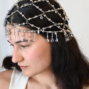 Pode incluir: Uma peça de cabeça prateada com um design de rede, adornada com pequenas contas e pingentes de cristal transparente. A peça é usada em cabelos escuros, criando um visual delicado e elegante. Perfeito para ocasiões especiais.