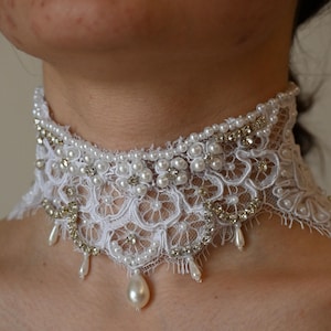 Pode incluir: Um colar gargantilha de renda branca adornado com pérolas e strass. O design intrincado apresenta padrões florais e detalhes de pérolas penduradas. Esta joia elegante é adequada para ocasiões especiais.
