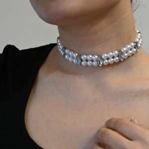 Puede incluir: Un collar gargantilla de perlas de doble hebra con detalles de cristal transparente. El collar está compuesto por perlas blancas y redondas y cristales transparentes de forma ovalada. El collar se lleva alrededor del cuello.