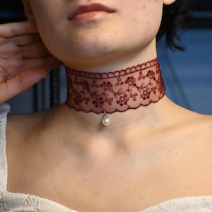 Puede incluir: Un collar gargantilla de encaje rojo con un delicado patrón floral y un colgante de perla.