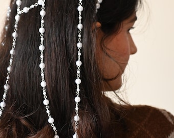 May include: A delicate headpiece featuring white pearl beads and silver-toned chains. The headpiece is designed to be worn across the forehead and cascades down the hair with dangling strands of pearls. The woman is wearing a brown and white patterned sweater.