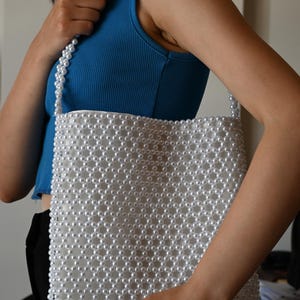 May include: A white beaded tote bag with a matching beaded strap. The bag is rectangular and densely covered in small, round, white beads. The strap is also made of white beads. The person in the image is wearing a blue tank top.