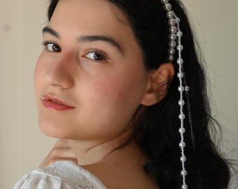 May include: A woman with dark hair is wearing a white pearl headband with long strands of pearls hanging down the back of her head. She is wearing a white, lacy, off-the-shoulder top.