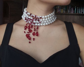 May include: A white pearl choker necklace with a dangling strand of red beads. The necklace is layered and has a delicate, feminine look.