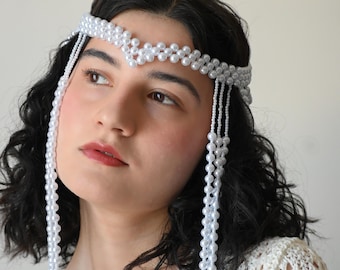 May include: A white beaded headband with dangling strands of beads. The headband is made of small, round, white beads and is worn on the head. The model is wearing a white crocheted top.